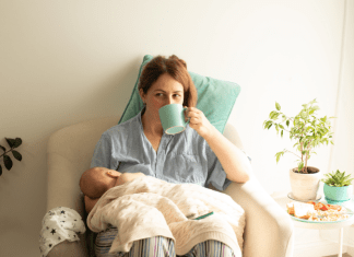 Mother is breastfeeding while sipping coffee, and there is a plate of food on her nighstand.