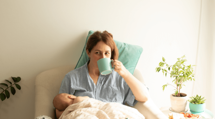 Mother is breastfeeding while sipping coffee, and there is a plate of food on her nighstand.