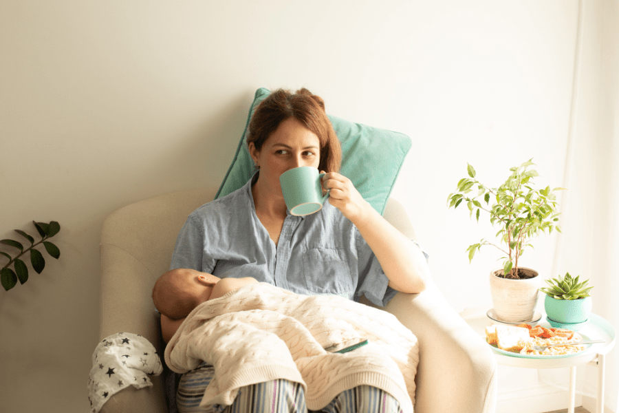 Mother is breastfeeding while sipping coffee, and there is a plate of food on her nighstand.