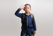 Kindergarten boy waves a salute goodbye as he wears a backpack and heads to school.