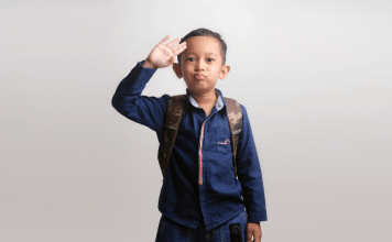 Kindergarten boy waves a salute goodbye as he wears a backpack and heads to school.