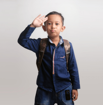 Kindergarten boy waves a salute goodbye as he wears a backpack and heads to school.