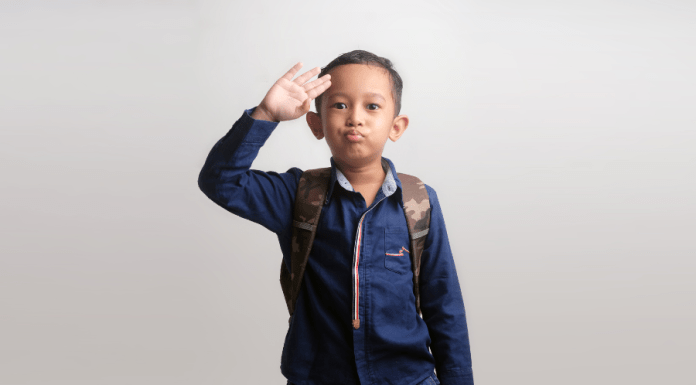 Kindergarten boy waves a salute goodbye as he wears a backpack and heads to school.