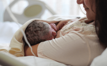 New mom looks lovingly at breastfeeding newborn while laying in hospital bed.