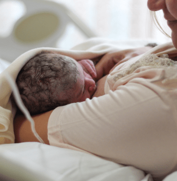 New mom looks lovingly at breastfeeding newborn while laying in hospital bed.