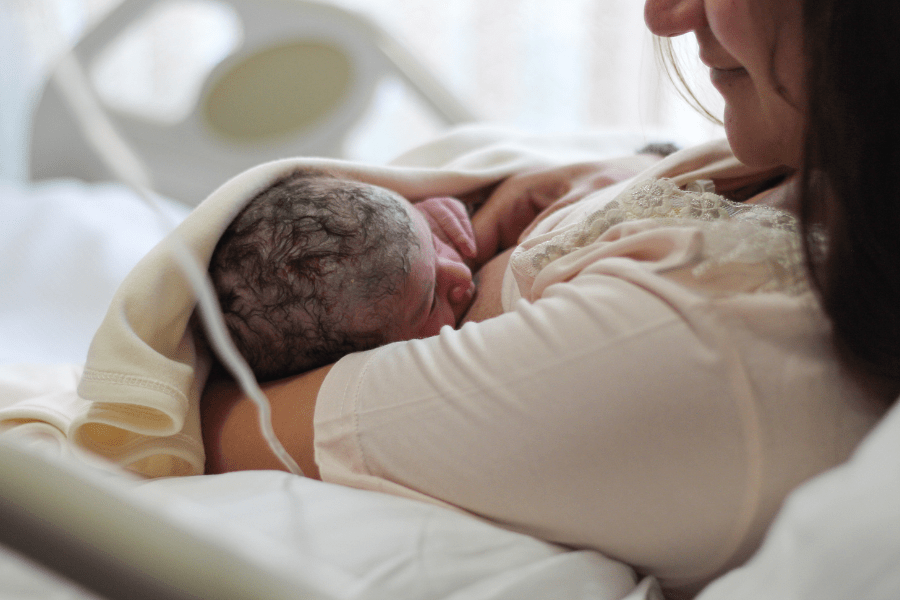 New mom looks lovingly at breastfeeding newborn while laying in hospital bed.