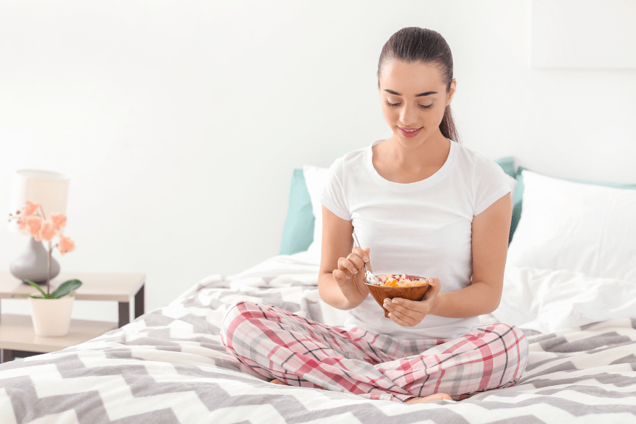 Mom sits cross-legged in bed, and eats a bowl of oatmeal.