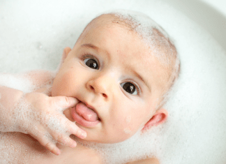 Baby lays in a soapy bath with a finger in his mouth, and looks surprised.