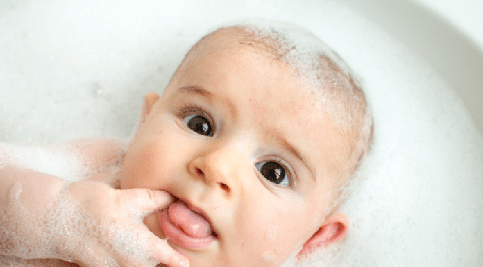 Baby lays in a soapy bath with a finger in his mouth, and looks surprised.