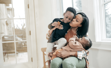 Tired mama holds a baby on her lap and her toddler in her arms as she kisses and loves on them.