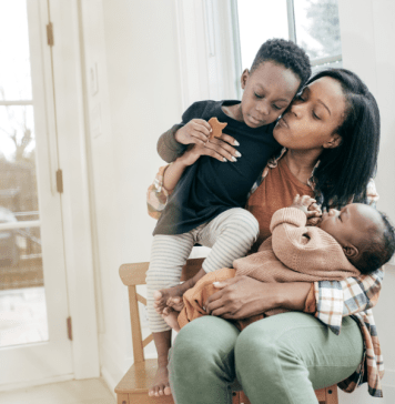 Tired mama holds a baby on her lap and her toddler in her arms as she kisses and loves on them.