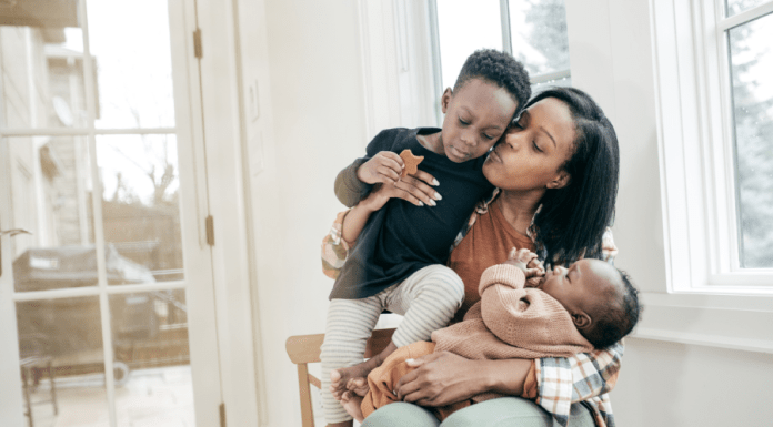 Tired mama holds a baby on her lap and her toddler in her arms as she kisses and loves on them.