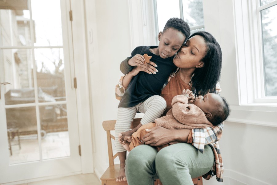 Tired mama holds a baby on her lap and her toddler in her arms as she kisses and loves on them.