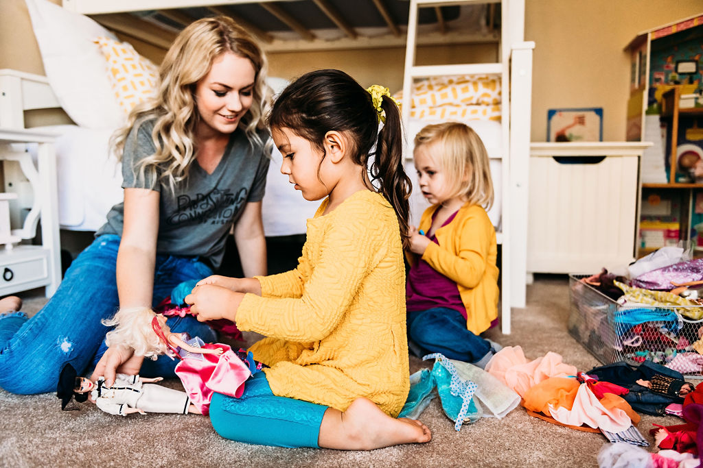 a babysitter sits on the floor with two children as they play with dolls