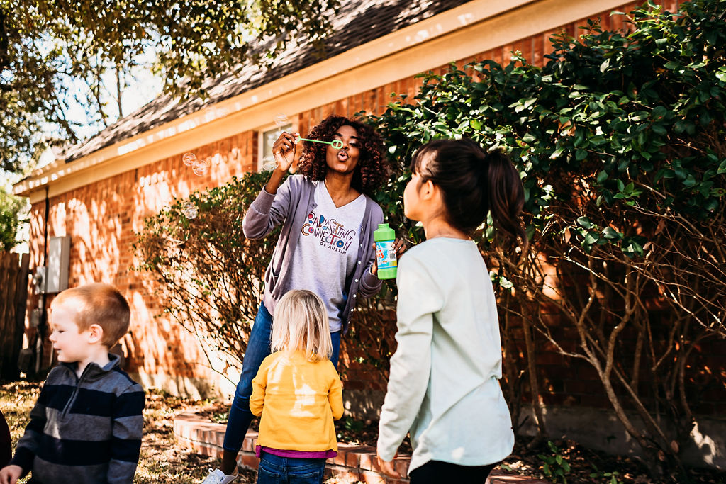 babysitter blows bubbles in back yard as young kids watch