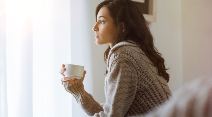 A woman holds a cup of coffee, looking out a window and thinking.