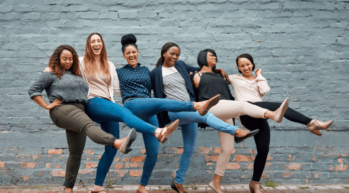 Six women of all types of skin colors locking arms and kicking one leg in the air, laughing