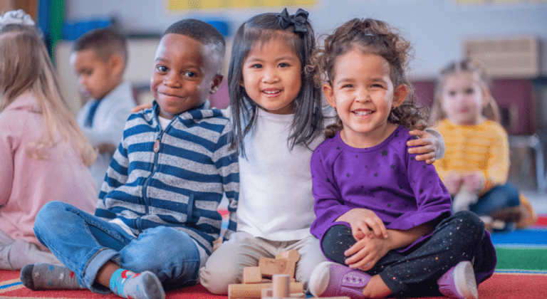 Three preschoolers sit on the ground with the middle child embracing them.