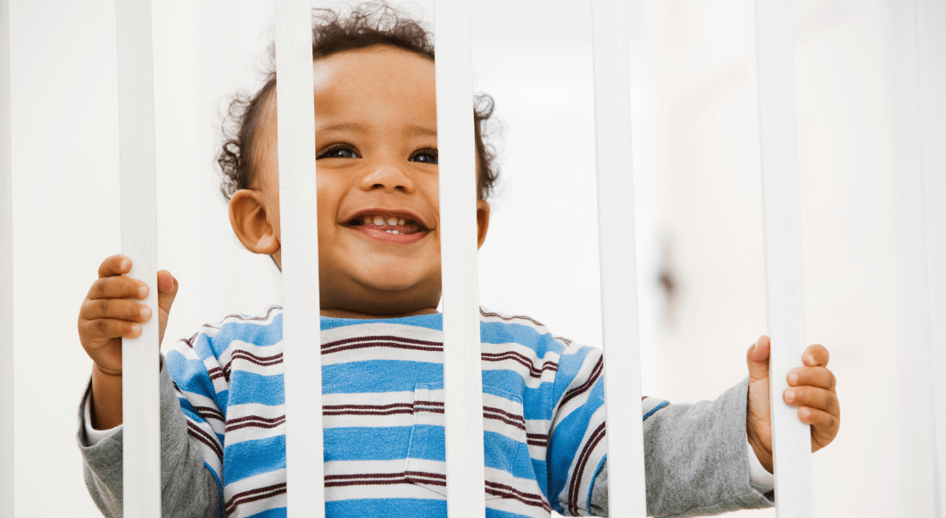 A baby stands behind a white babygate.