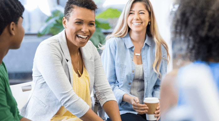 A group of moms sit in a circle laughing and drinking coffee.