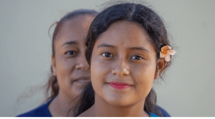 A Latina mom stands behind her daughter, who has a flower in her hair.