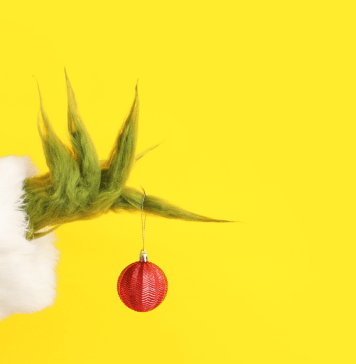 The Grinch's hand holding a red ornament with a yellow background