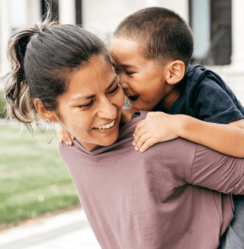 Little boy rides piggyback on his mom's back in front of a house.