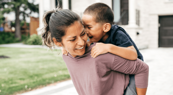 Little boy rides piggyback on his mom's back in front of a house.