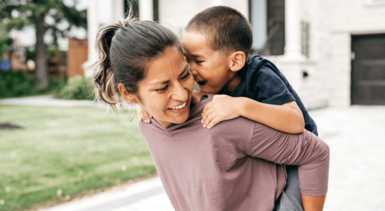 Little boy rides piggyback on his mom's back in front of a house.