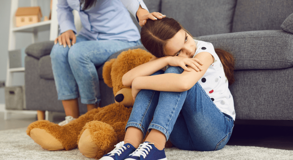 A little girl hugs her knees and feels sad, while her mom strokes her hair.