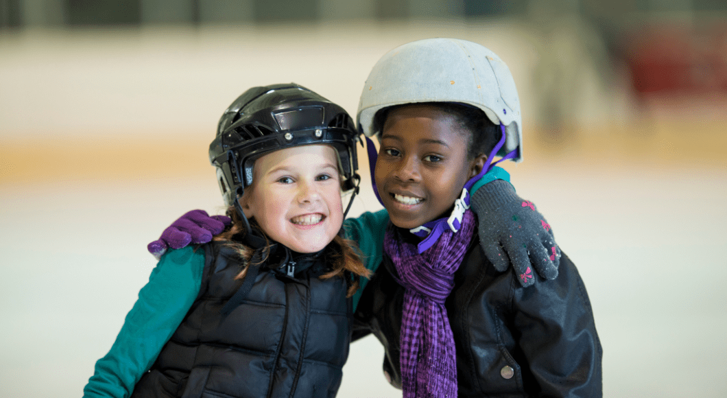 Two girls smile together at indoor ice skating rink.