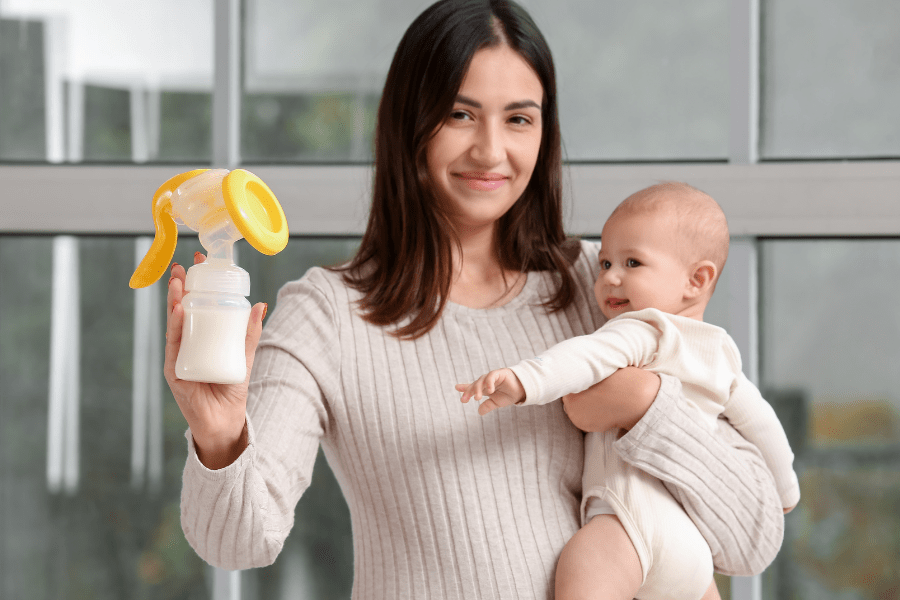 Mother holds baby in one arm and a breast pump bottle in the other, and smiles while looking at the camera.