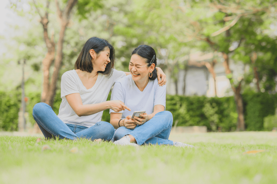 Mom mentor and mentee laugh together while sitting in a park.