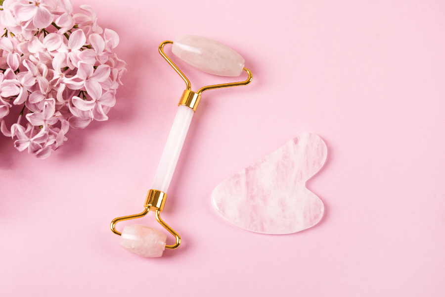 Pink flowers, rose quartz face roller, and a rose gua sha massager sit on a pink background.