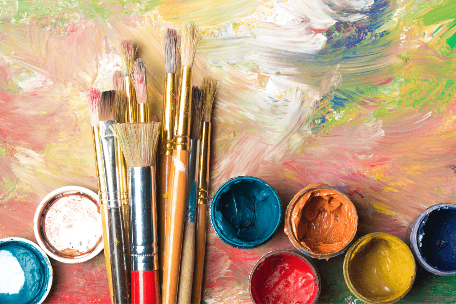 Paint brushes and pots of paint lay on top of an abstract painting.